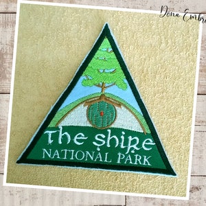 Könnte beinhalten: Grüner dreieckiger gestickter Aufnäher mit einem Baum, einer runden Tür und dem Text "The Shire National Park".