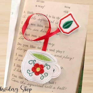 May include: A red ribbon bookmark with a white teacup and a green leaf design. The teacup has a red flower with a green stem. The bookmark is on a book with the text "A little squirrel is in the" and "now old".