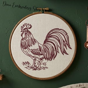 Puede incluir: Un bastidor bordado a mano con un diseño de gallo. El gallo está representado en un contorno detallado de color burdeos sobre una tela color crema. El bastidor de madera es redondo y tiene un cierre de latón. El texto "Dona Embroidery Shop" está en la parte superior.