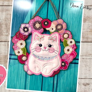 Puede incluir: Un gato rosa y blanco con un lazo en la cabeza está sentado en el centro de una corona de flores. La corona está hecha de flores de tela rosa, blanca y verde. El fondo es una superficie de madera turquesa. El texto "Instructions Included" está en un corazón rosa en la esquina inferior izquierda.