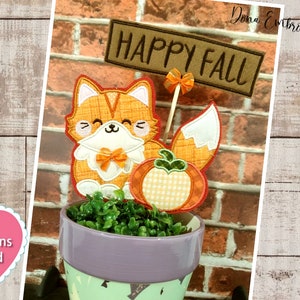 May include: Embroidered applique design featuring a cute orange fox holding a pumpkin with a bow. The fox is sitting in a pot with green plants and a sign that says "Happy Fall".