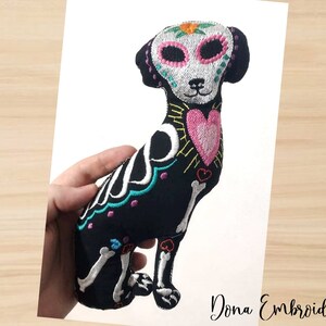 May include: A black plush dog with embroidered details in pink, white, blue, and green. The dog is depicted as a skeleton with a heart on its chest and a flower on its head. The dog is sitting up with its legs crossed. "Dona Embroidery Shop" is written in the bottom right corner of the image.