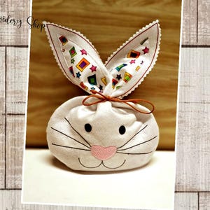 May include: White fabric bunny-shaped gift bag with colorful patterned ears. The bag has a pink nose and embroidered whiskers.