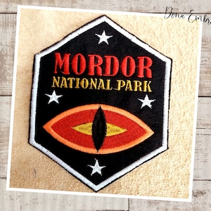 May include: Black embroidered patch with a white outline. The patch features a red eye with a yellow pupil and the text "Mordor National Park" in white and red lettering.