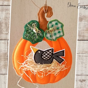 May include: A felt pumpkin wreath with a bird in a nest, green leaves, and a checkered pattern. The wreath is orange and brown with a loop for hanging. "Dona Embroidery Shop" is written in the top right corner.