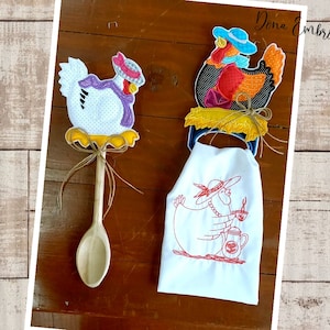 Può includere: Due porta-asciugamani da cucina ricamati con galline che indossano cappelli. Un porta-asciugamani è un cucchiaio di legno con una gallina bianca che indossa una sciarpa viola e un cappello rosa. L'altro porta-asciugamani è un asciugamano bianco con una gallina rossa che indossa un cappello blu e tiene una tazza di caffè. Il testo "Dona Embroidery Shop" è visibile nell'angolo in alto a destra dell'immagine.