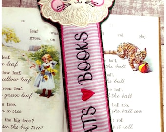 Peut inclure: Un marque-page rayé rose et blanc avec une tête de chat brodée en blanc et le texte "CATS BOOKS" en noir. Un cœur rouge est brodé entre les mots "CATS" et "BOOKS".