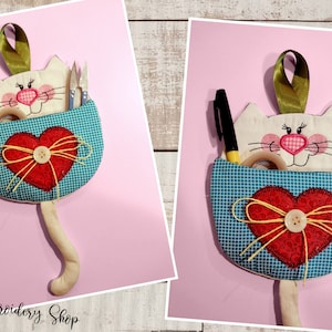 Może przedstawiać: Biały organizer z tkaniny w kształcie kota z kieszenią w niebiesko-białą kratę, czerwonym sercem z żółtą kokardką i żółtą wstążką zwisającą z góry. Organizer jest wypełniony różnymi materiałami do rękodzieła, w tym nożyczkami, długopisem i taśmą klejącą.