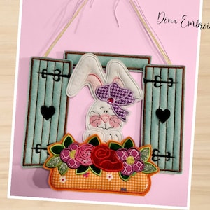 Puede incluir: Un conejito blanco con un lazo morado se sienta en una ventana con contraventanas azules. El conejito está rodeado de flores rosas, rojas y amarillas en una maceta. La ventana está decorada con corazones negros. El texto "Dona Embroidery Shop" es visible en la esquina superior derecha de la imagen.