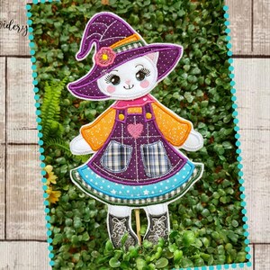 Pode incluir: Uma aplicação caprichosa em forma de gato com um chapéu de bruxa, mangas laranja e um vestido xadrez roxo. O design inclui um coração rosa e está sobre um fundo de vegetação e uma superfície de madeira. O texto "Dona Embroidery" está no canto superior esquerdo.