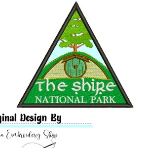 Op de afbeelding: Een groene driehoekige patch met een witte rand. De patch toont een groene heuvel met een ronde bruine deur en een groene boom. De tekst "The Shire National Park" is in wit gedrukt.