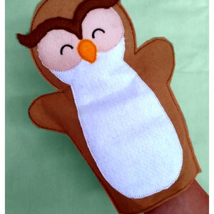 May include: A brown felt owl hand puppet with a white chest, brown wings, and a smiling face. The owl has dark brown accents, an orange beak, and black stitched eyes. The puppet is designed to fit over a hand.