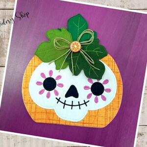 以下が含まれることがあります： 砂糖の頭蓋骨の顔、ピンクの花、そして上にリボンが付いた緑の葉を持つフェルトの南瓜。南瓜はオレンジと白で、背景は紫です。