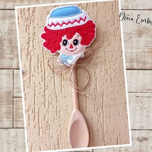 May include: A wooden spoon with a red-haired doll head attached to the top. The doll has a blue and white hat and a blue bow. The spoon is on a light brown wooden background.