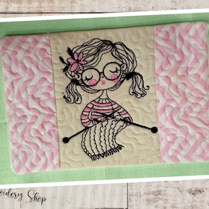 Könnte beinhalten: Ein gesteppter Untersetzer mit einem pink-weißen Zickzackmuster an den Seiten. Die Mitte zeigt ein gesticktes Design eines Mädchens mit Brille, das einen Schal strickt.