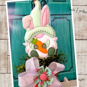 May include: A white bunny gnome with a green hat and a carrot in its hand. The gnome is sitting in a pot with a pink bow and green foliage.