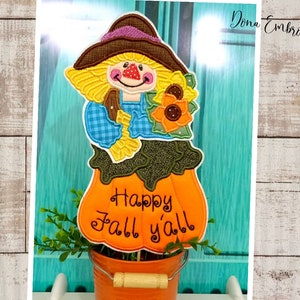 Pode incluir: Um espantalho bordado colorido com uma abóbora e girassóis, segurando um ancinho. A abóbora tem o texto "Happy Fall y'all". O espantalho está em um pequeno vaso laranja com uma alça de madeira.