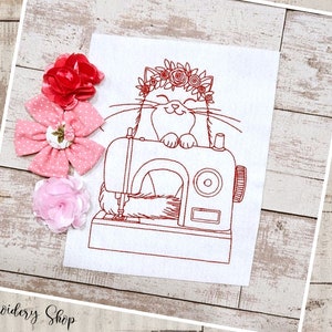 Pode incluir: Desenho de bordado de arte de linha de um gato usando uma coroa de flores sentado em uma máquina de costura. O desenho é em fio vermelho em tecido branco.