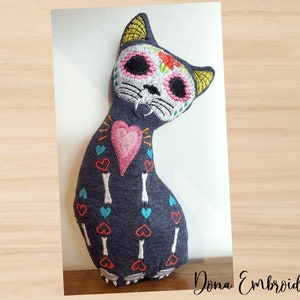May include: A gray fabric cat plush toy with embroidered details in black, white, pink, blue, yellow, and orange. The cat is decorated with a Day of the Dead inspired skull and crossbones design, hearts, and bones. The text "Dona Embroidery Shop" is visible in the bottom right corner of the image.