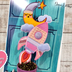 Pode incluir: Um foguete rosa e branco com um detalhe de glitter prateado, uma lua amarela com um rosto adormecido e nuvens. O foguete está em um palito e está na frente de uma porta azul turquesa. O texto "Instructions Included" está em um coração rosa.