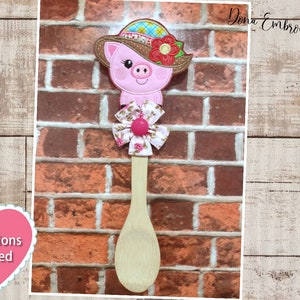 May include: A wooden spoon with a pink pig wearing a hat and a floral bow. The pig has a pink nose and pink ears. The spoon is on a brick background. Instructions included.