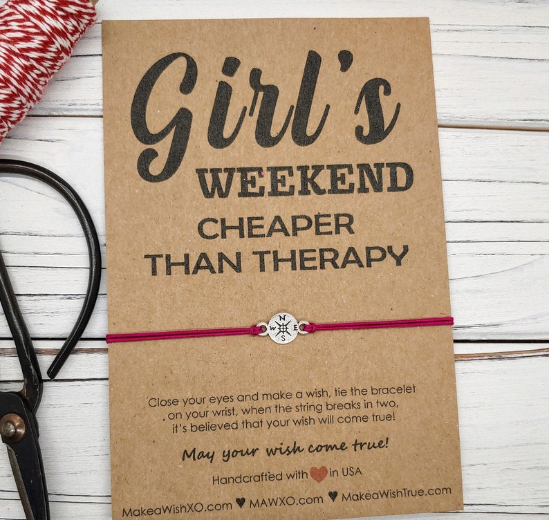 May include: A wish bracelet on a brown card with the text "Girl's Weekend Cheaper Than Therapy." The bracelet has a magenta string and a silver compass charm. The card also includes text about making a wish.