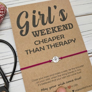 May include: A card with the text "Girl's Weekend Cheaper Than Therapy" and a magenta bracelet with a compass charm. A roll of red and white twine and scissors are also visible.