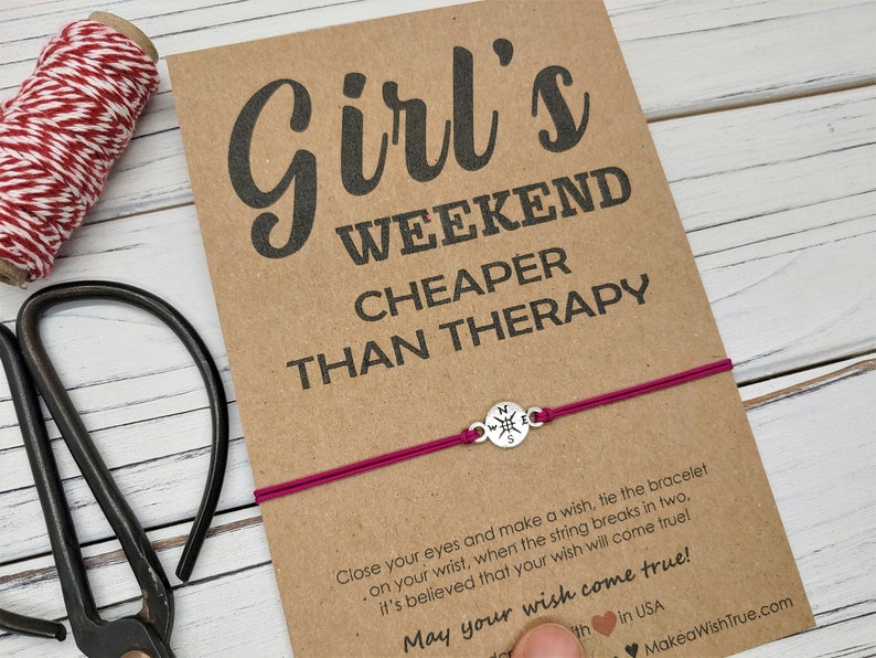 May include: A card with the text "Girl's Weekend Cheaper Than Therapy" and a magenta bracelet with a compass charm. A roll of red and white twine and scissors are also visible.
