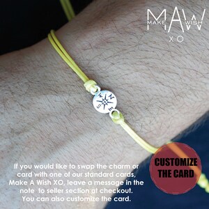 May include: A yellow cord bracelet with a silver compass charm. The bracelet is tied with knots and is shown on a wrist. The compass charm has the letters N, E, S, and W. The text "MAW MAKE WISH XO" is in the background.