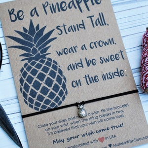 May include: A brown card with a black pineapple illustration and the text "Be a Pineapple, stand tall, wear a crown, and be sweet on the inside." A black bracelet with a small pineapple charm is attached.