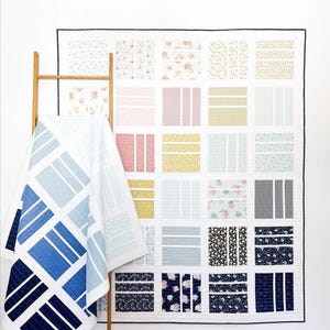 May include: A quilt with a patchwork design featuring various patterns and colors, including blue, white, pink, yellow, and gray. The quilt is hanging on a white wall and is folded in half.