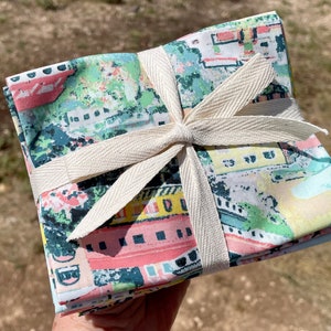 May include: A stack of colorful fabric squares with a city scene print, tied together with a natural-colored ribbon. The fabric is a mix of pink, blue, green, and yellow.