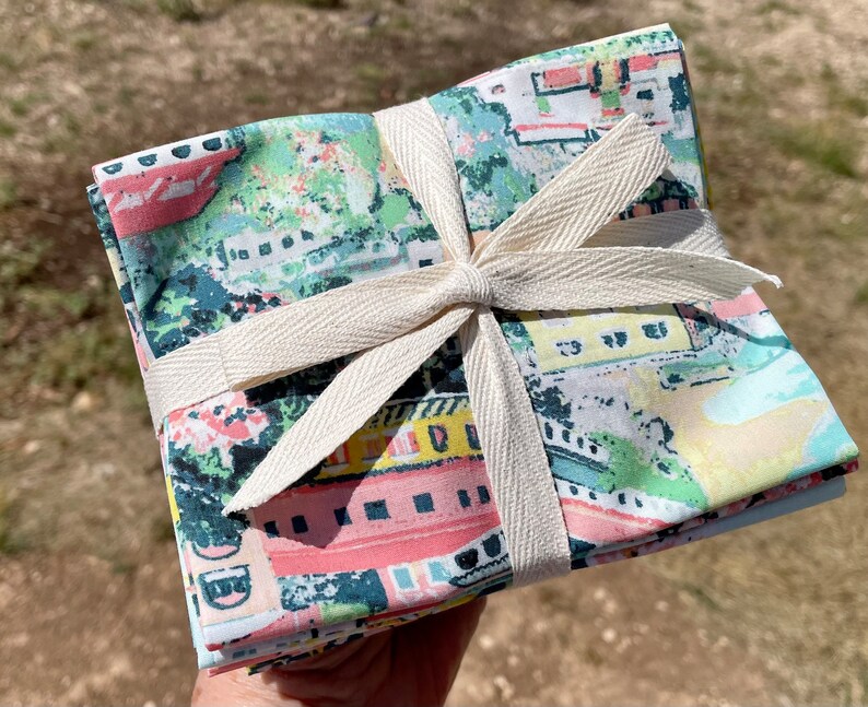 May include: A stack of colorful fabric squares with a city scene print, tied together with a natural-colored ribbon. The fabric is a mix of pink, blue, green, and yellow.