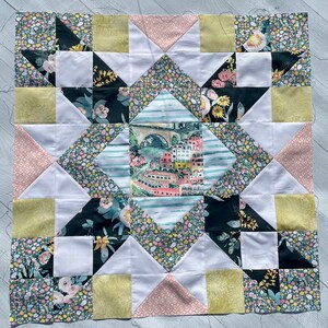 May include: A quilt block with a star design featuring a variety of floral and geometric patterns in shades of pink, green, yellow, and black. The center of the block features a watercolor illustration of a cityscape with a bridge.