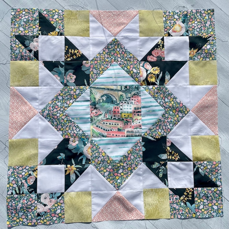 May include: A quilt block with a star design featuring a variety of floral and geometric patterns in shades of pink, green, yellow, and black. The center of the block features a watercolor illustration of a cityscape with a bridge.