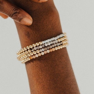 May include: A stack of beaded bracelets in gold, white, and pink tones. The bracelets are worn on a person's wrist.