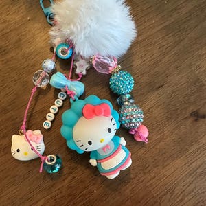 May include: A colorful keychain featuring Hello Kitty charms and a fluffy white pom-pom. The keychain includes a blue bow, beaded accents, and the word "CUTIE" spelled out in white letter beads. The Hello Kitty charms are in blue, pink, and white.