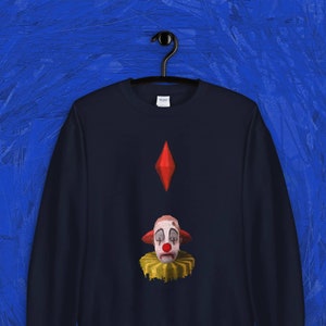Puede incluir: Una sudadera azul marino con un diamante rojo y un gráfico de cara de payaso triste. El payaso tiene un cuello amarillo y una nariz roja.