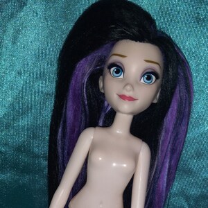 May include: A nude doll with long black and purple hair. The doll has blue eyes and a painted-on smile. The doll is standing on a teal fabric background.