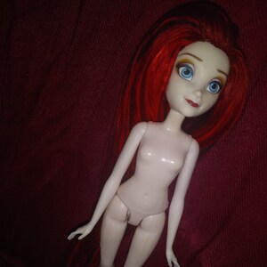 May include: A nude doll with long red hair and blue eyes. The doll is posed with its legs spread and its arms raised.