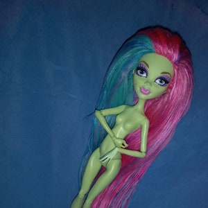 Peut inclure: Une poupée à la peau verte avec de longs cheveux roses et bleus. La poupée porte un corsage blanc et est posée avec une main sur la hanche et l'autre main près de son visage.