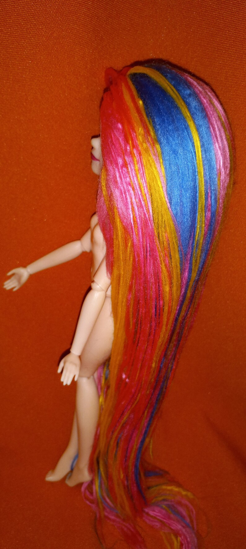 May include: A doll with long, colorful hair. The hair is pink, yellow, blue, and orange. The doll is nude and has a simple face.
