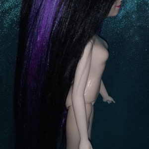 May include: A doll with long black hair with purple streaks. The doll is wearing a nude-colored outfit and has a light blue background.