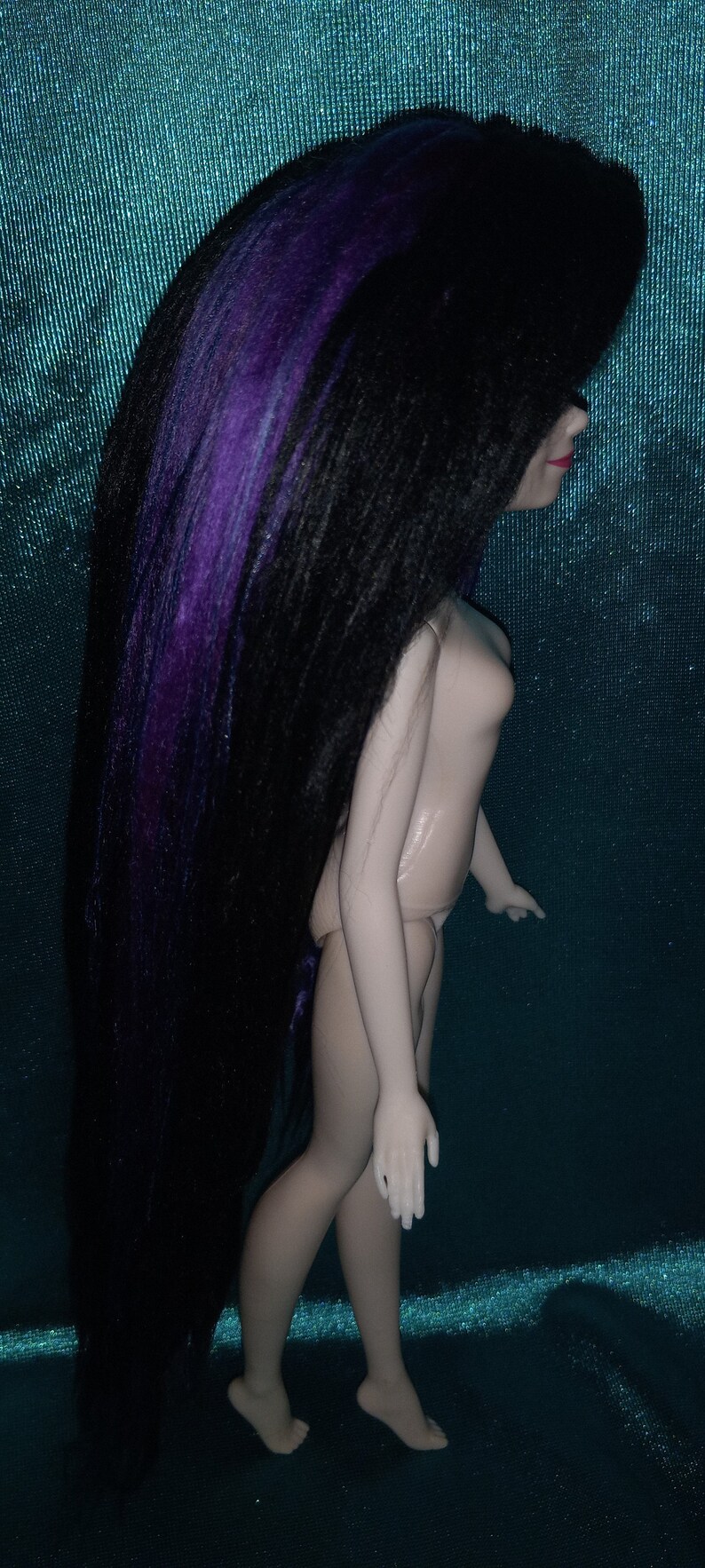 May include: A doll with long black hair with purple streaks. The doll is wearing a nude-colored outfit and has a light blue background.