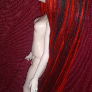 May include: A doll with long, red and black dreadlocks. The doll is nude and has a white, porcelain-like body. The doll is posed with one leg bent and the other leg straight. The doll is standing on a red fabric background.