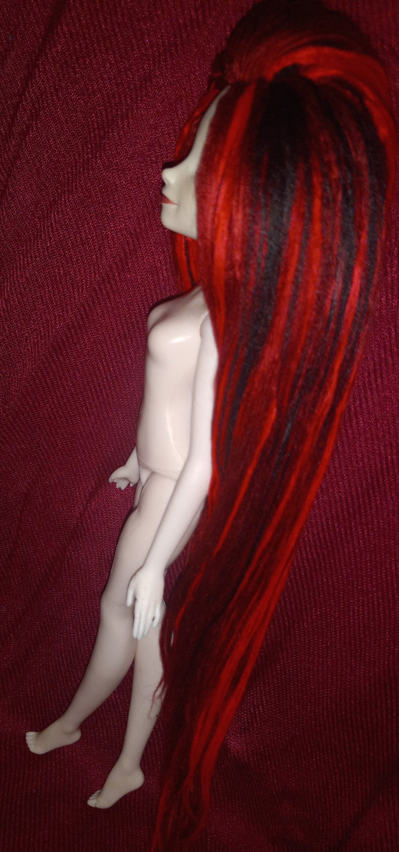 May include: A doll with long, red and black dreadlocks. The doll is nude and has a white, porcelain-like body. The doll is posed with one leg bent and the other leg straight. The doll is standing on a red fabric background.