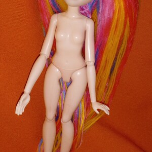 May include: A nude doll with long, colorful yarn hair. The hair is pink, yellow, blue, and green. The doll has blue eyes and a painted smile.