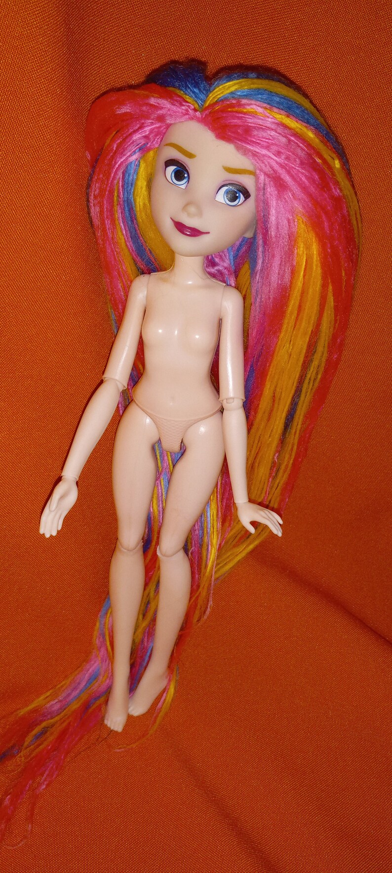 May include: A nude doll with long, colorful yarn hair. The hair is pink, yellow, blue, and green. The doll has blue eyes and a painted smile.