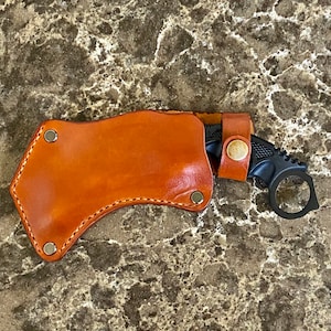 May include: A black karambit knife with a finger ring, partially sheathed in a brown leather holster. The holster is secured with metal rivets and a leather strap with a snap closure. The knife and holster are on a speckled surface.