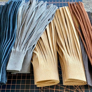 May include: Five strips of leather fringe in various colors, including black, gray, beige, brown, and dark brown. The fringe is cut into long, thin strips and is arranged in a row on a black and orange cutting mat.